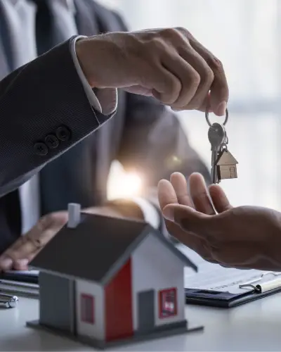 Agent handing house keys to a buyer beside a model house